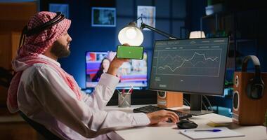 Arab man watching video on green screen cellphone while looking at business annual report charts on computer. Worker spending time on chroma key mobile phone while solving job tasks at home photo
