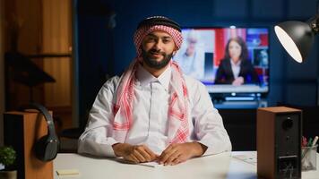 Cheerful Arab man paying attention in elearning seminar teleconference with teacher. Middle Eastern student in online videocall greeting tutor, listening to lesson while writing down homework tasks photo