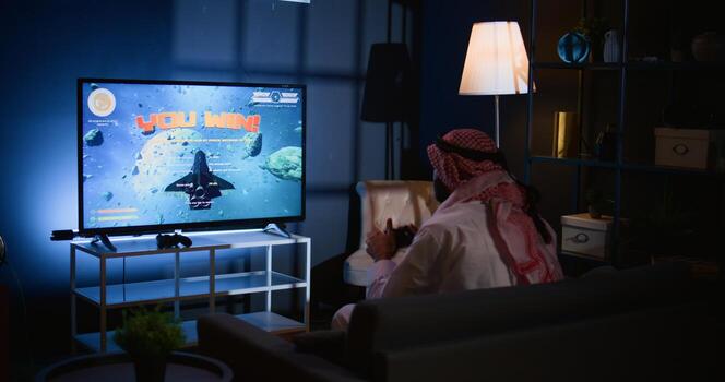 Middle Eastern gamer plays intense classic arcade space shooter videogame. Man relaxing at home using high tech gaming system handheld controller to solve missions in singleplayer game photo