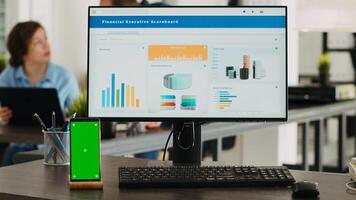 vacío oficina puesto de trabajo con pantalla verde corriendo en teléfono inteligente mostrar, personas trabajando en negocio operaciones en agencia lugar de trabajo. escritorio con computadora y móvil teléfono demostración aislado espacio de copia. foto