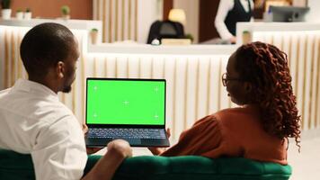 Happy couple on their honeymoon vacation holding laptop chroma key green screen mock up, talking with family in video call. Husband and wife chatting in video conference sitting in hotel lobby photo