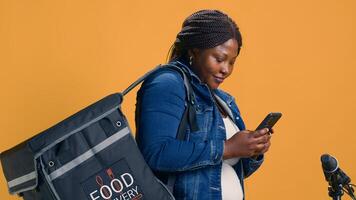 antes de montando para comida entrega Servicio un entusiasta entrega mujer usos su teléfono inteligente a mensaje compañeros de trabajo africano americano mensajero exploración un móvil aplicación para parcelas a entregar Bajo demanda. foto