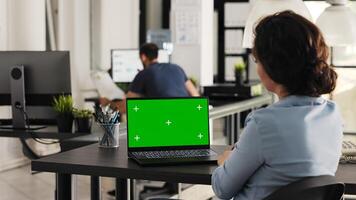 Businesswoman works on laptop with greenscreen, looking at blank chromakey display in coworking space. Young adult examining pc screen with isolated mockup template, e commerce tasks. Handheld shot. photo