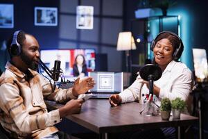 Female influencer laughing during comedy talk show filmed with smartphone on tripod stand. Young guest amused by talking with journalist telling jokes, recording with mobile device. photo