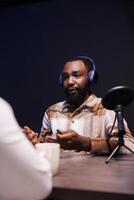 negro masculino anfitrión entrevistando un persona a hogar, utilizando moderno micrófono y inalámbrico auriculares para su en línea hablar espectáculo. africano americano hombre hablando con un individual grabación un podcast episodio. foto