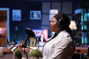 Side view portrait of african american female talk show host speaking with her online audience during a live podcast recording from her home studio, she wears wireless headphones. photo