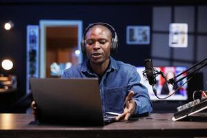 Black male influencer creates material for his internet audience, discussing new trends. African American man wearing headphones is speaking into a microphone and filming a promotional video. photo