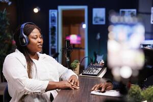 durante el entrevista, un negro hembra celebridad registros contenido para su en línea audiencia desde su hogar estudio utilizando un teléfono inteligente en un trípode mientras respondiendo preguntas desde un entrevistador. foto