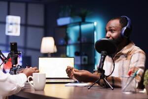 Young black man with a notebook is giving advice online using podcast equipment. At the table is an african american male vlogger speaking into the microphone, recording an episode at home. photo