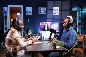 Sitting at table using the audio equipment are two african american men discussing and addressing their online audience. Interviewer asking male influencer questions, recording a radio show at home. photo