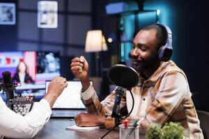 Smiling black man doing fist bump with african american presenter before starting live radio broadcast. Cheerful male influencer greeting guest during a recording session of online talk show. photo