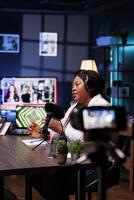 Black woman creates vlogs using professional camera while producing podcasts with audio equipment. Female journalist interviews guests in her home studio, filming with a device on a tripod. photo