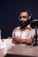 African american man using audio equipment to create an online radio show at home. Young black male content creator with wireless headphones and speaking into a microphone, is making a podcast. photo