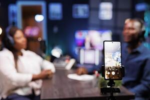 A vlogger is recording a podcast episode with a smartphone mounted on a tripod in a neon-lit studio. The show host is interviewing a guest and filming the discussion using a mobile device. photo