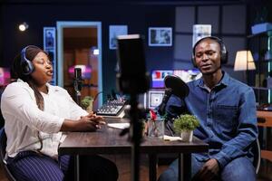 African American content creator couple talks tech in their home studio, entertaining a wide online audience with engaging discussions. Black vloggers using mobile device for recording radio program. photo