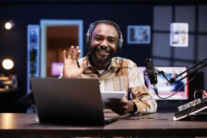 Young male vlogger waving towards the camera, filming channel intro using recording equipment at home. African american content creator doing a live broadcast, making viral video for online audience. photo