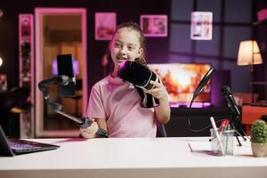 Kid excited to review VR headset, comparing features with older models, filming with smartphone on selfie stick. Cute girl analyzing high tech virtual reality goggles, recording with phone camera photo