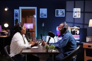 Cheerful african american couple having a conversation at home, and using audio equipment for podcast episode. Joyful male and female influencers wearing wireless headphones and talking into mics. photo