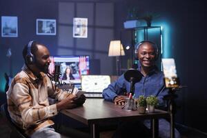 During live debate on vlog channel, happy vloggers utilizing smartphone on a tripod to film podcasts. Joyful men gathered in a studio to record broadcast using a cell phone and discuss world affairs. photo