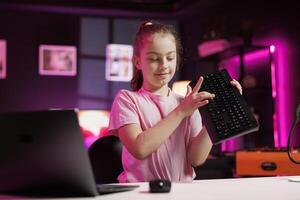Happy charismatic girl presenting latest gaming computer peripheral tech on her internet channel in neon lit apartment. Trending influencer kid filming review of wireless Bluetooth mechanical keyboard photo