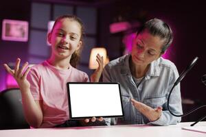 Girl and mother film reaction video for gen Z subscribers, looking on mockup tablet at online videos. Child media star and her parent watching funny internet clips on isolated screen device photo