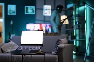 Focus on isolated screen laptop on coffee table in apartment with man in blurry background cleaning dust on shelves. Close up shot of mockup notebook and person in the back with tablet in hand photo