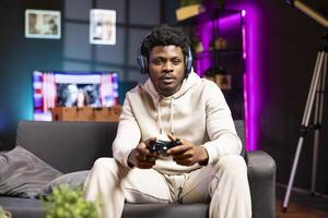 Man sitting on couch at home, relaxing by playing videogames on gaming console. BIPOC gamer unwinding in living room by competing in online multiplayer esports tournament photo