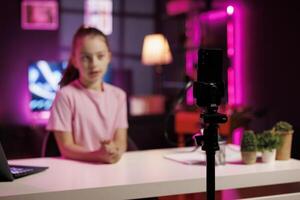 Focus on cellphone on tripod used by kid shooting product unboxing for streaming channel on online platforms. Child in blurry background warmly welcoming fans, filming reviews with mobile phone photo