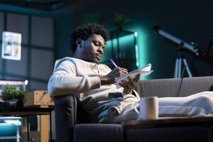 Entrepreneur working from home, writing business ideas on notebook. African american man lounged on couch using pen to note plan on paper while teleworking in cozy living room photo