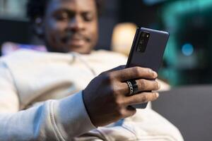 Close up of cellphone held by person chatting with online friends, enjoying himself at home. Focus on mobile phone used by BIPOC man in blurry background to scroll on social media photo