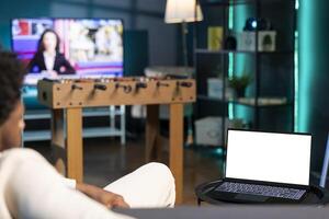 Man relaxing after work in apartment by watching TV series on mockup laptop. Person in neon illuminated home looking at shows on isolated screen notebook with television set left open on news channel photo