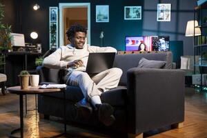 Cheerful african american man checking emails on laptop while laying on couch. Happy BIPOC freelancer working from home in bright stylish cozy apartment personal office, using notebook device photo