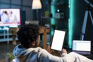 Man sitting on couch, reading eBook from tablet in apartment using mockup tablet. Relaxed person enjoying interesting lecture on isolated screen device at home, relaxing after work photo