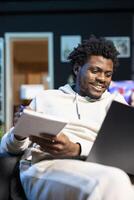 Entrepreneur working from neon lit apartment, writing down company ideas while watching video on laptop. BIPOC man feeling delighted after coming up with startup proposal, using pen to note plan photo