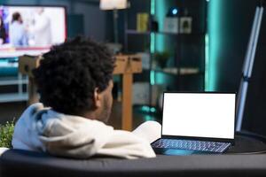 Remote employee inputting data on mockup laptop display while working from home, looking at screen. Teleworker lying on couch, remotely performing tasks on isolated screen notebook in living room photo