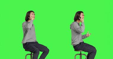 Modern guy making phone calls on camera, sitting on chair against greenscreen backdrop and having remote conversation. Young person being relaxed talking to friends, telephone line. photo