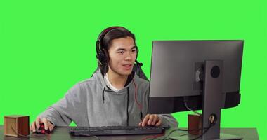 joven persona sentado a escritorio oficina, teniendo divertido con amigos mientras disfrutando en línea multijugador juegos con auriculares. terminado pantalla verde, asiático chico compite en papel jugando concurso. foto