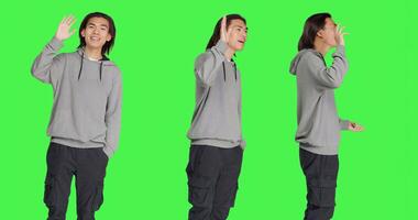 Asian guy waving hello at someone, greeting people with a smile and confidence against greenscreen template. Young adult saying hi with a wave, smiling and feeling cheerful in studio. photo