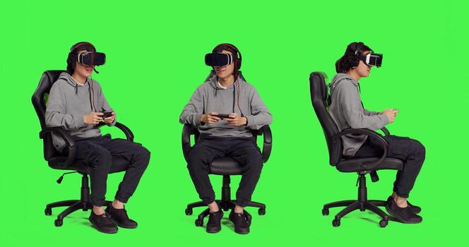 Person rests on chair over greenscreen backdrop and performs video games using virtual reality eyewear and mobile device, takes part in online multiplayer contest. Asian guy loving combat games. photo