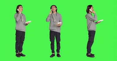 Asian guy talking on landline phone, answering call on telephone with cord while he stands against full body greenscreen. Young person using retro office phone for remote communication. photo
