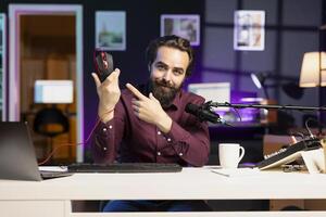 Smiling influencer filming video for online channel in apartment studio, presenting gaming mouse to audience. Happy content creator showing viewers computer peripheral device, endorsing it photo