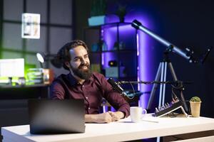 Happy internet star using quality mic in studio to discussing with viewers. Smiling man sitting at table in front of laptop using high tech recording gear to record social media video photo