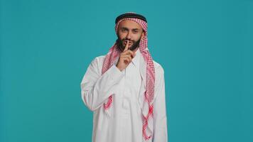 Arab man does silence sign with finger over lips, showing hush mute gesture in studio. Private model presenting confidential secrecy symbol on camera, trying to keep secret and privacy. photo