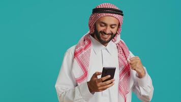 Arab guy holding his mobile device, surfing across social media platforms and chatting online. Muslim with customary apparel uses smartphone to access global web, feeling excited on camera. photo