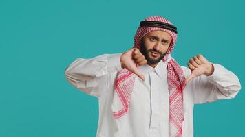 Muslim person gives thumbs down and feeling displeased about something, indicating arabic culture with traditional robe and headscarf. Young adult showing refusal and negative gesture. photo