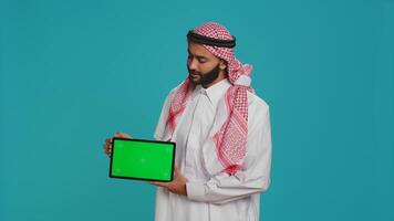 Muslim man presents greenscreen tablet over blue background, showing blank copyspace display. Young person in arabic clothing holding gadget with isolated chromakey mockup template. photo