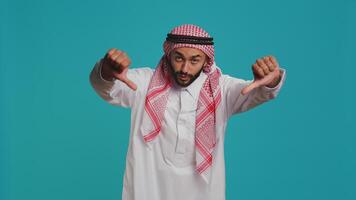 Arab adult with national gown and kufiyah shows thumbs down and feels dissatisfied with news. Young man expresses an apparent denial and negative gesture on camera, arabic person. photo