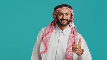 Confident man showing like sign in studio, giving thumbs up and agreement while he represents traditional arabic culture. Middle eastern positive guy wears islamic costume and headscarf. photo