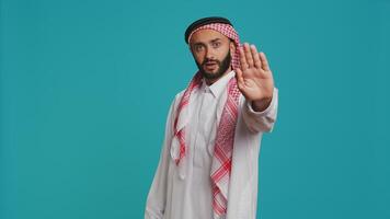 Middle eastern person does stop sign with palm up, expressing refusal and denial on studio camera. Young man presenting rejection symbol, being displeased and disappointed. photo