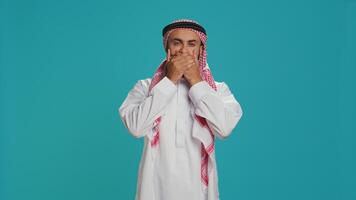 Muslim adult covers eyes, ears and mouth in studio, showcasing three wise monkeys metaphor sign. Middle eastern guy presenting dont hear, see or speak concept symbol in arabic clothes. photo